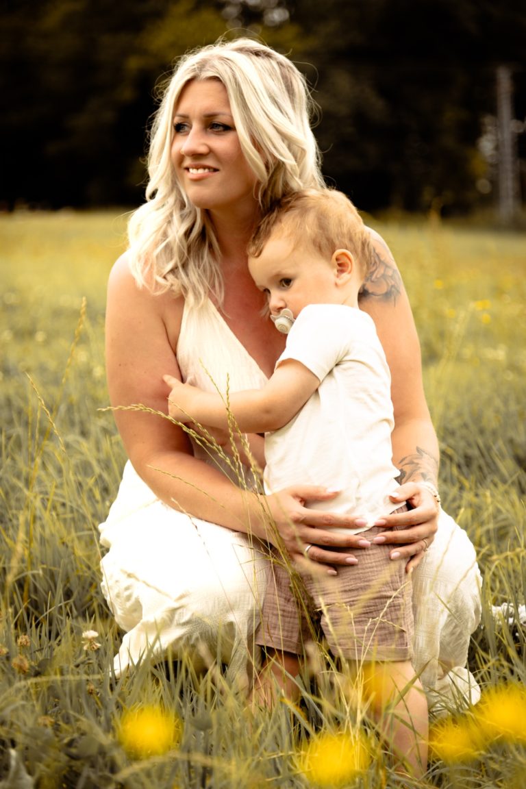 Frau mit blonden Haaren umarmt ein Kleinkind in einem blühenden Feld.