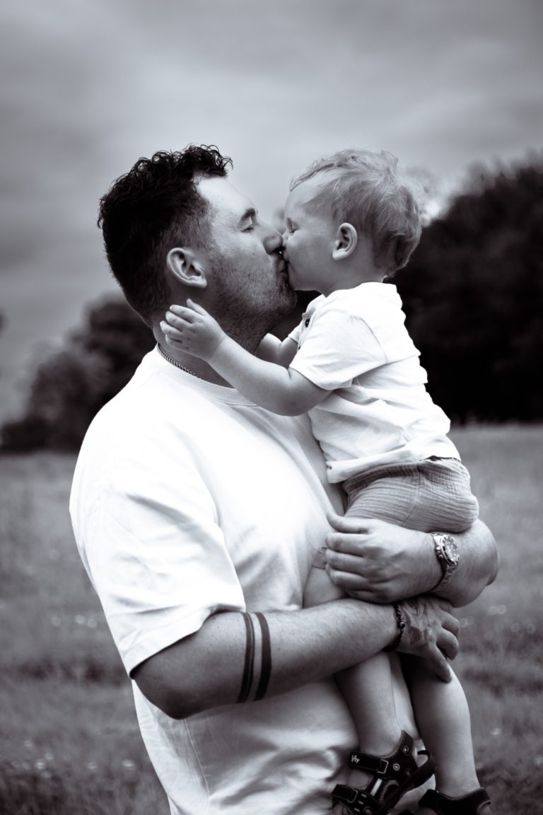 Vater küsst sein Kleinkind in einer grasbewachsenen Landschaft.