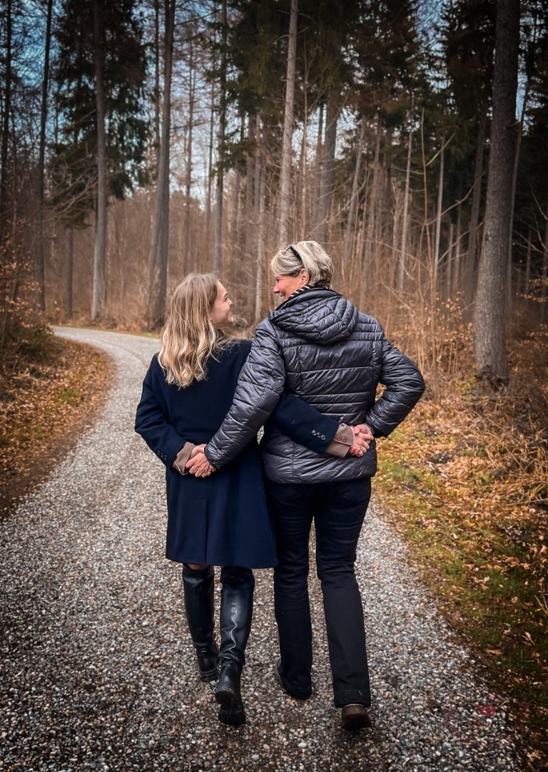 Zwei Frauen gehen Hand in Hand einen Waldweg entlang, Rücken zur Kamera.