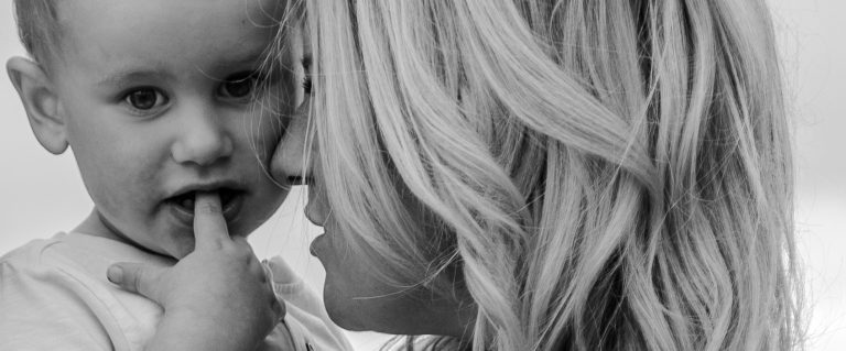 Eine Frau umarmt ein Kind, das neugierig mit dem Finger in den Mund greift.