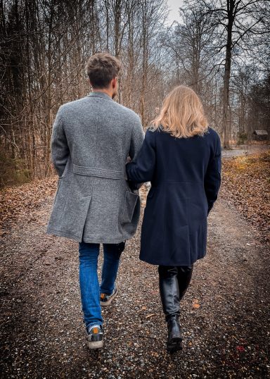 Ein Paar geht Hand in Hand einen Waldweg entlang, umgeben von Bäumen und Laub.