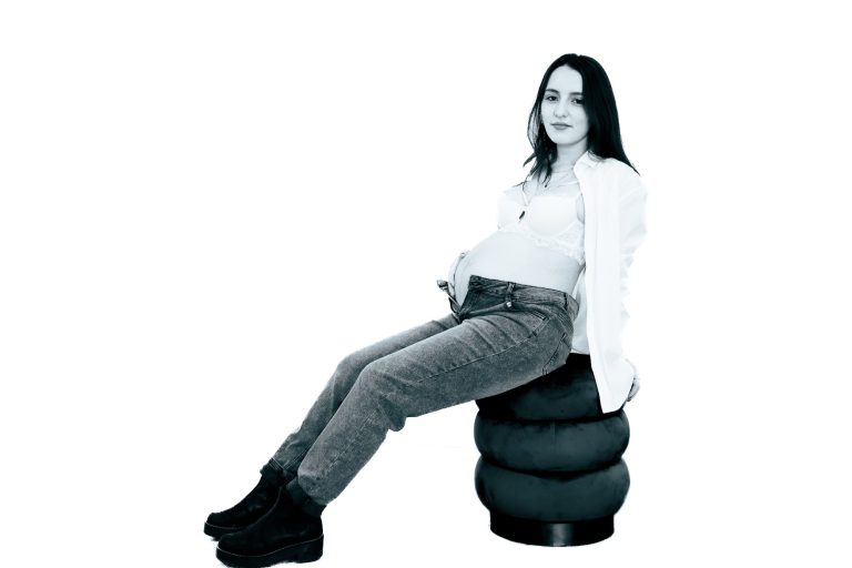 Eine Person sitzt auf einem runden Hocker, trägt eine weiße Bluse und Jeans.