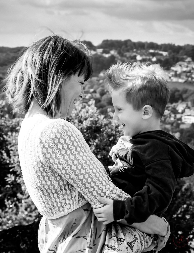 Eine Frau hält einen Jungen in ihren Armen, beide lächeln sich an, mit Landschaft im Hintergrund.