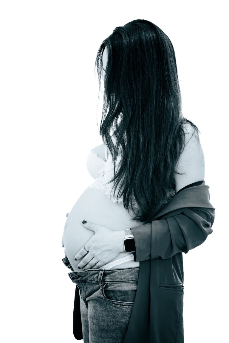 Schwangere Frau hält ihren Bauch, mit langen, dunklen Haaren und lässiger Kleidung.