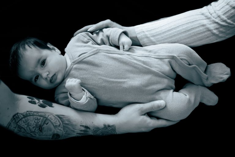 Ein Baby in einem grauen Overall, gehalten von zwei Händen, eine mit Tattoo.