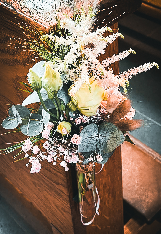 Ein bunter Strauß aus Rosen, Eukalyptus und Trockenblumen, an einer Holzbank befestigt.