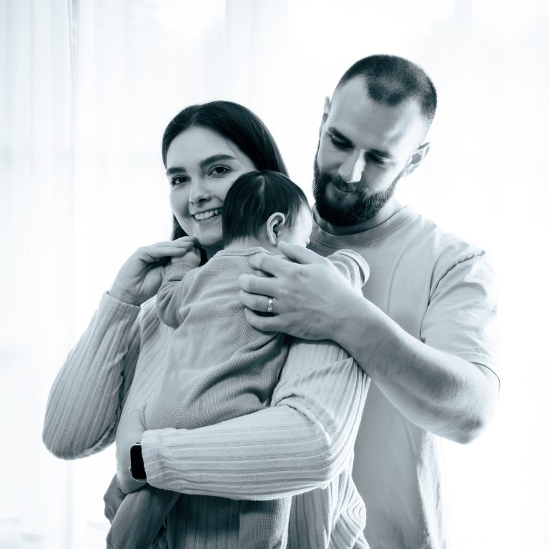Eine Familie umarmt ihr Baby, lächelnd in einer hellen Umgebung.