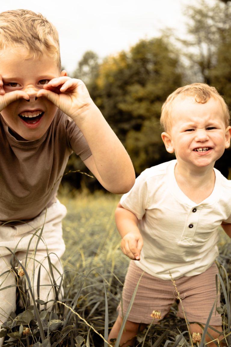Zwei kleine Jungen stehen im Gras, einer lächelt und hält die Hände vor den Augen.