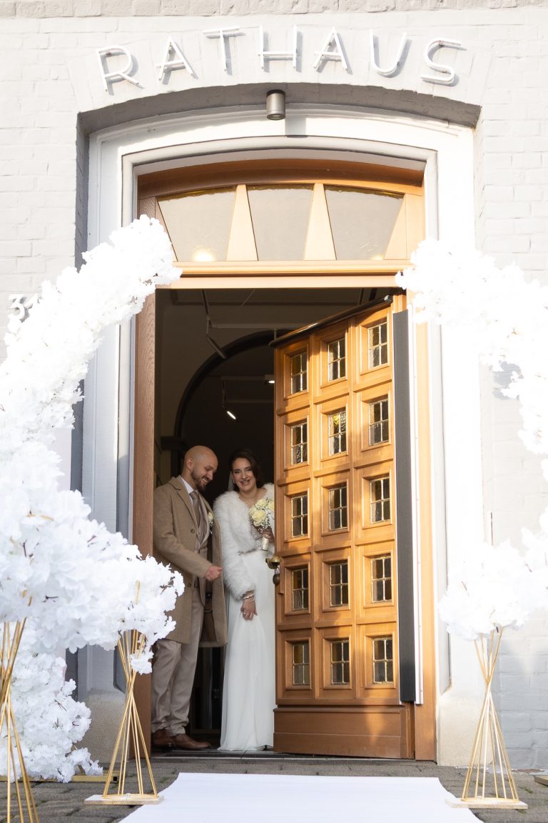 Brautpaar tritt in festlicher Kleidung vor das Rathaus mit dekorativen weißen Elementen.