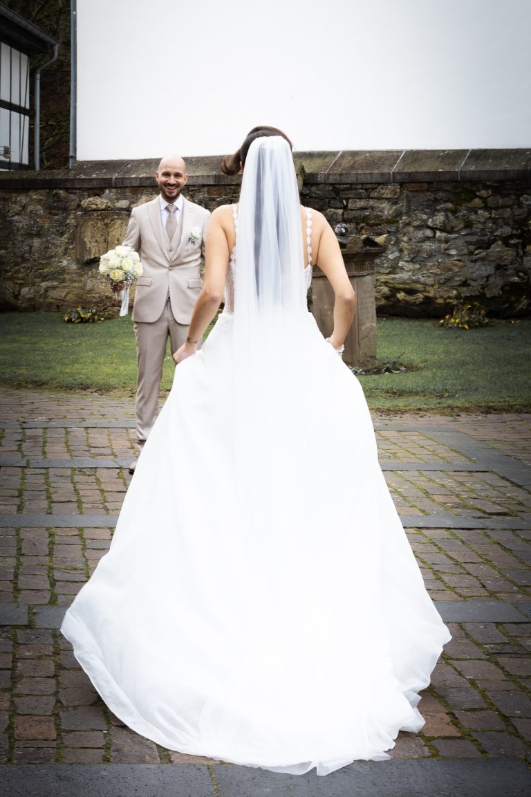 Braut in weißem Kleid mit Schleier blickt den Bräutigam an. Hintergrund mit Steinmauer.