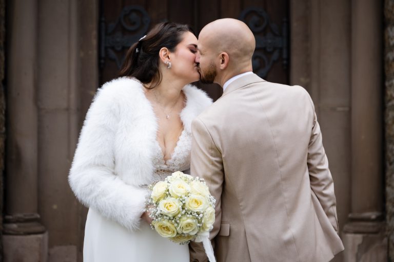 Paar in Hochzeitskleidung küsst sich vor einer historischer Tür, Braut hält einen Blumeneinsatz.