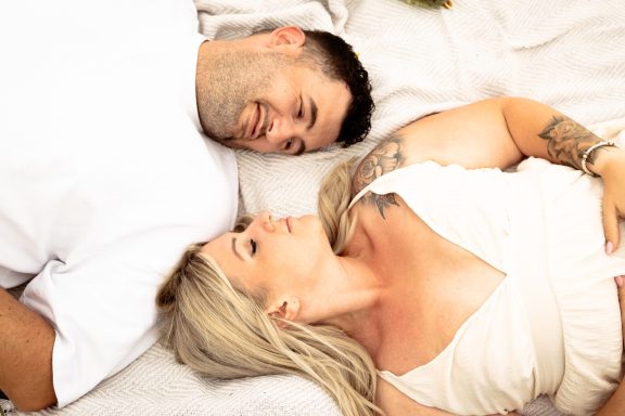 Paar liegt entspannt nebeneinander auf einer Decke, beide lächeln sich an.