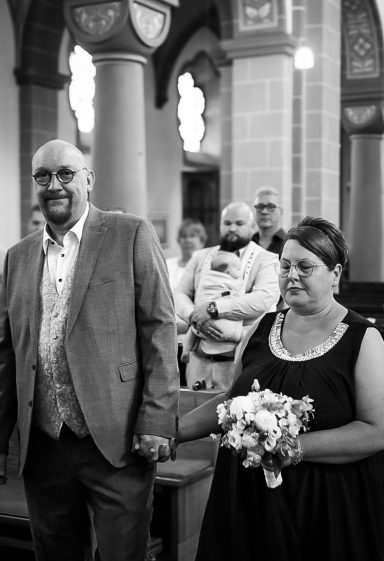 Hochzeitspaar geht gemeinsam den Gang entlang, Gäste im Hintergrund beobachten sie.