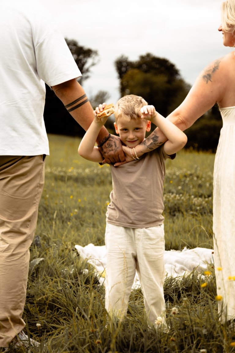 Ein Kind steht zwischen zwei Erwachsenen in einem Feld, umarmt von ihnen und lächelt.