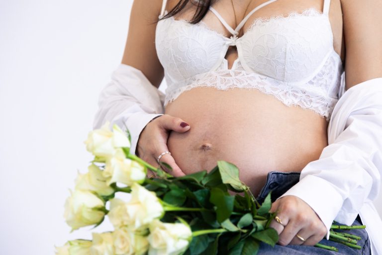 Schwangere Frau in Unterwäsche hält ihren Bauch und eine Vase mit weißen Rosen.