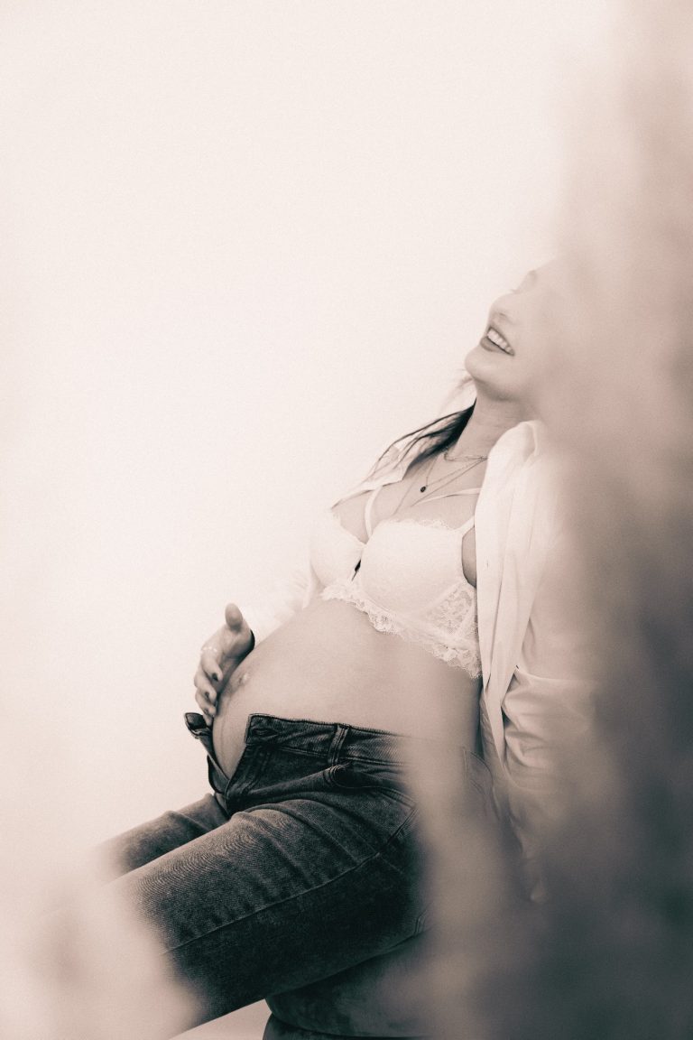 Schwangere Frau sitzt entspannt und hält ihren Bauch, sanfte Farben im Hintergrund.