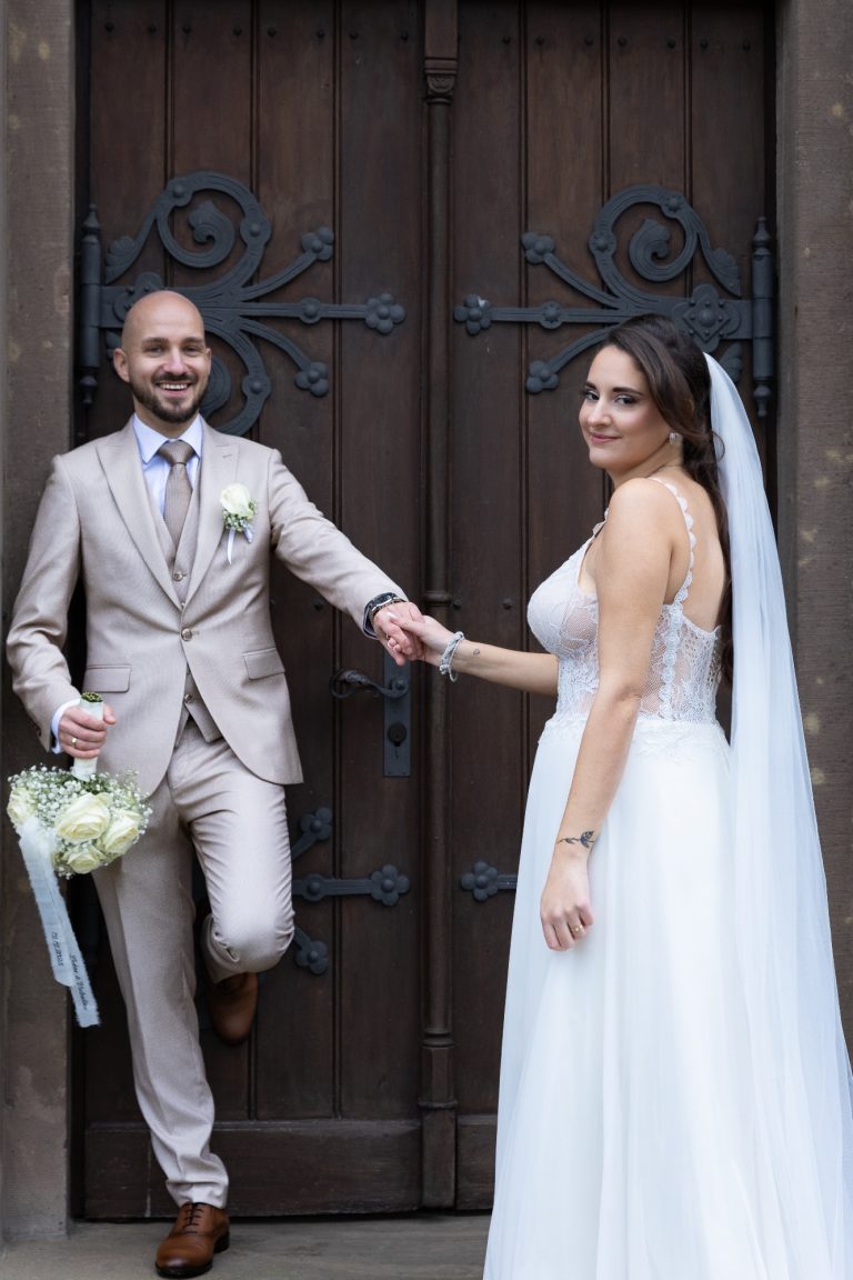 Braut und Bräutigam stehen vor einer Holzür, lächelnd, mit Blumenstrauß in der Hand.
