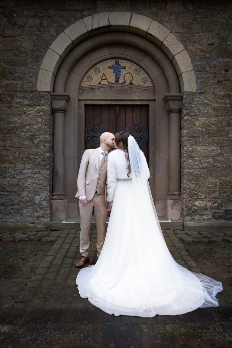 Braut und Bräutigam küssen sich vor einer steinernen Kirche im Freien.