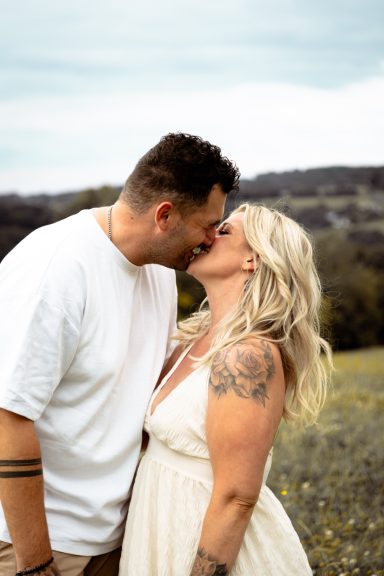 Paar küsst sich in einer blühenden Landschaft, umgeben von sanften Hügeln.