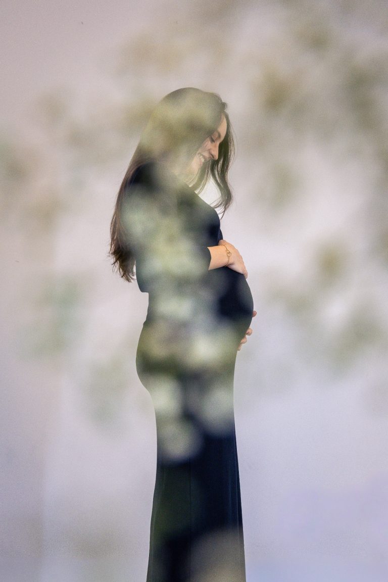 Schwangere Frau in schwarzem Kleid, sanft umarmt sie ihren Bauch, unscharfer Hintergrund.