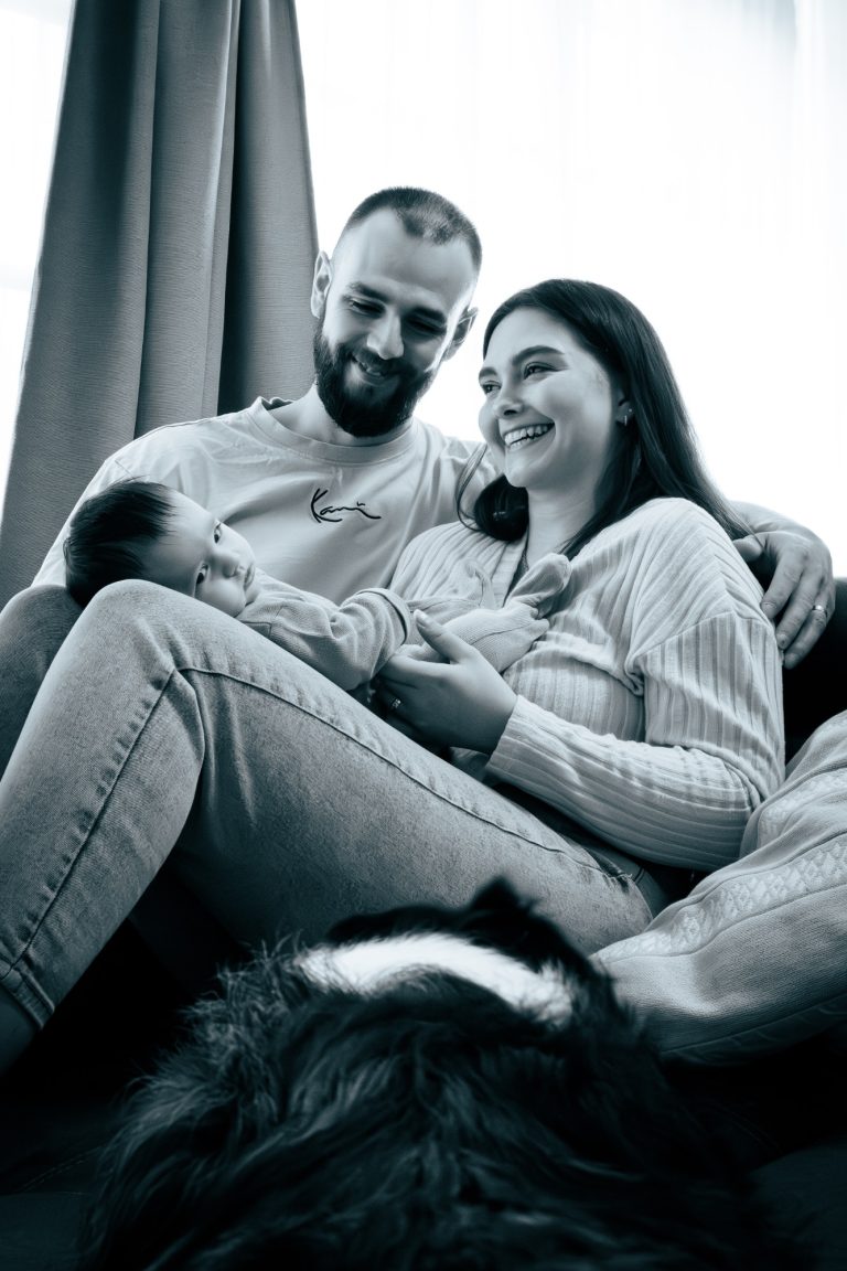 Ein Paar sitzt auf einem Sofa und hält ein Baby, ein Hund liegt entspannt davor.